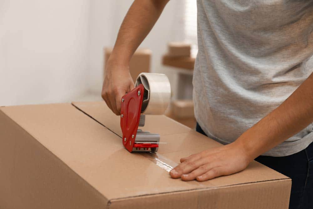 Man In Grey Shirt Packing Items Using A Tape Dispenser